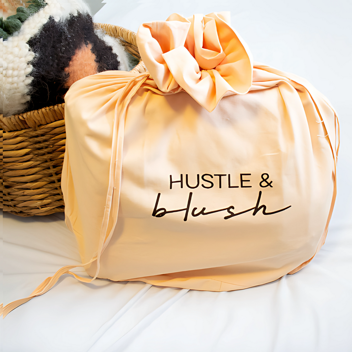Peach-colored fabric bag with "Hustle & blush" text beside a woven basket containing the Faino (Fine) Wool Statement Throw / Baby Blanket, displayed on a white surface.