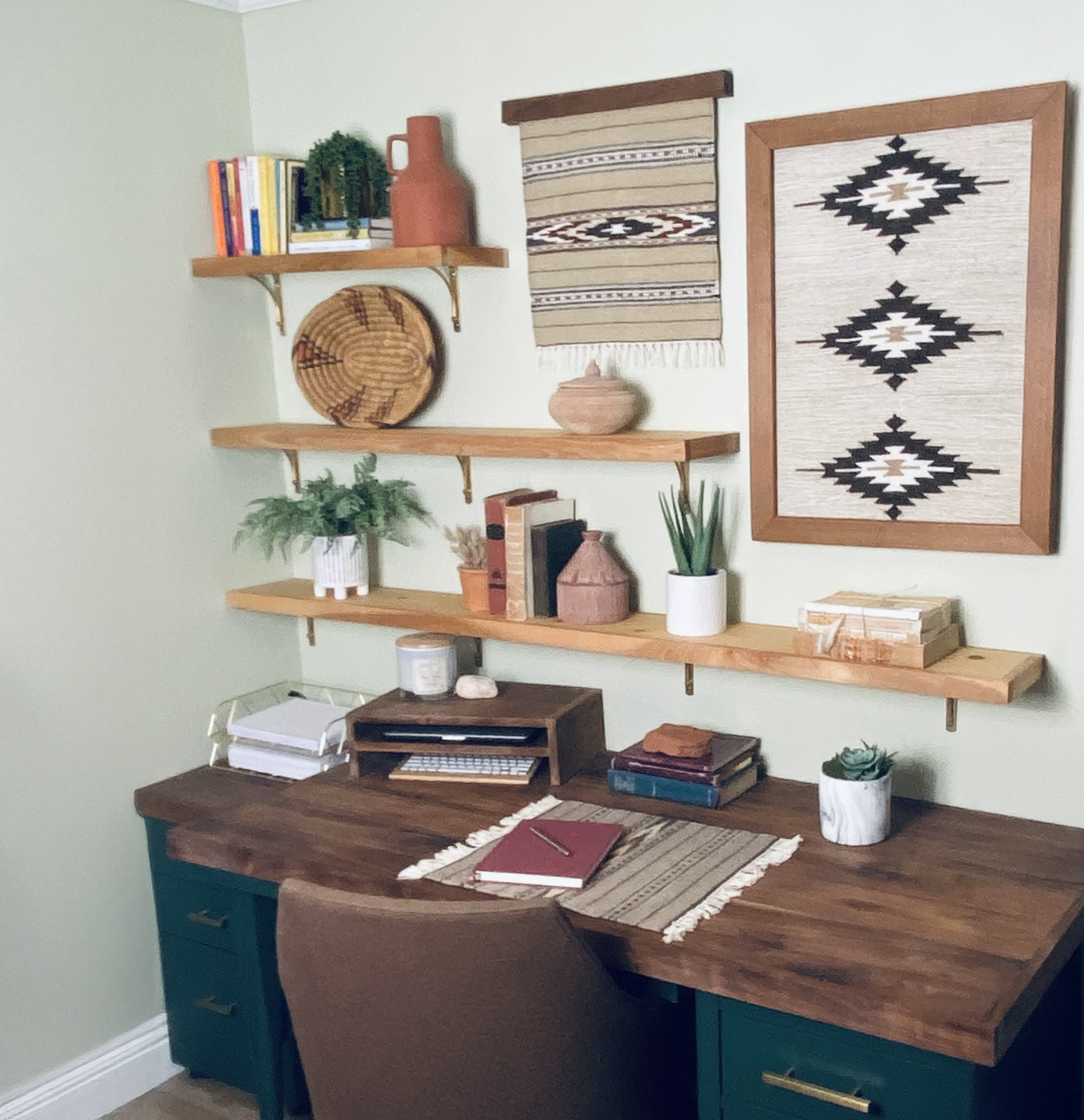 A tidy home office with wooden shelves, books, plants, and Southwestern woven decor above a wooden desk features the Wool Rug Framed Wall Art, adding warmth and sustainable style to the space.