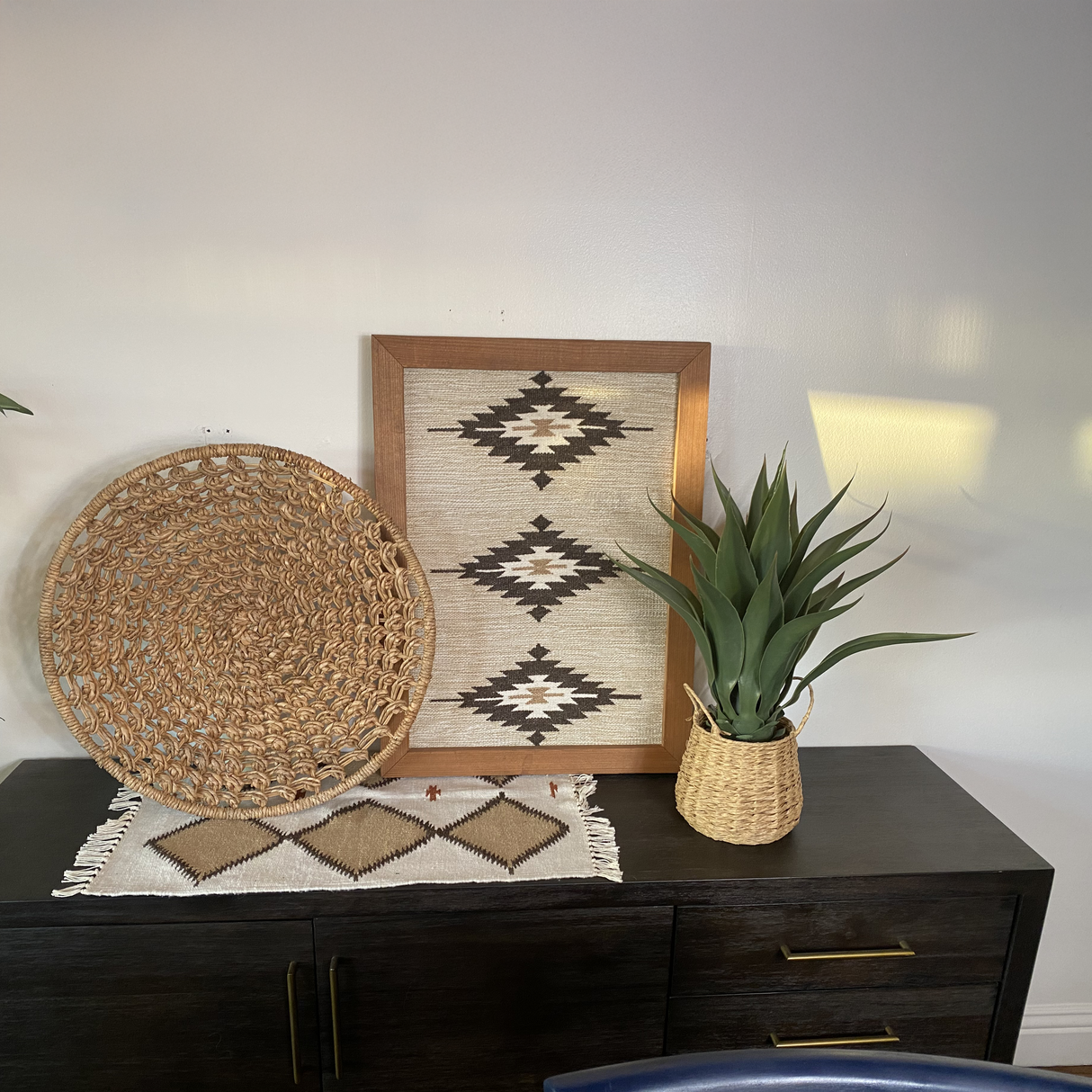 A woven basket and potted plant sit on a dark western cedar cabinet, beneath Wool Rug Framed Wall Art, all set against a white wall.