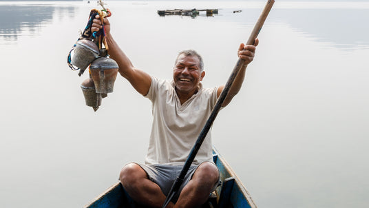 A smiling man sits in a boat, holding up metal bells, with calm water in the background.