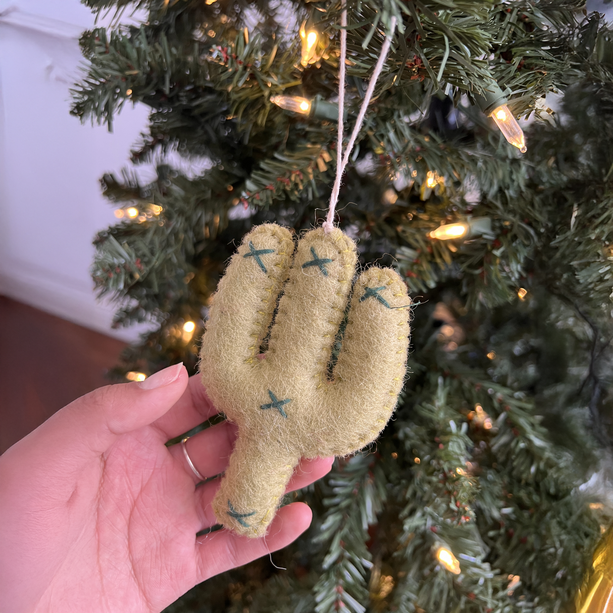 A hand displays the Hand-Stitched Felted Cactus Ornament in front of a glowing Christmas tree, highlighting the unique charm of handmade felt decor.