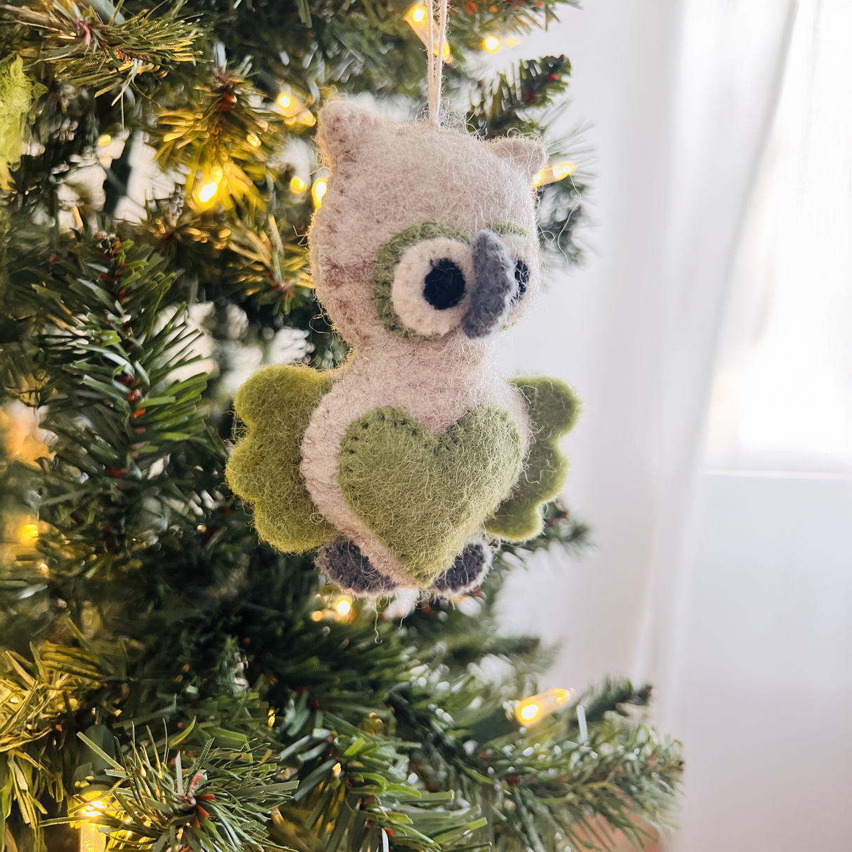 The Hand-Stitched Felted Owl Ornament with green wings and a heart hangs on a lit Christmas tree, bringing charm to your artisan holiday décor.