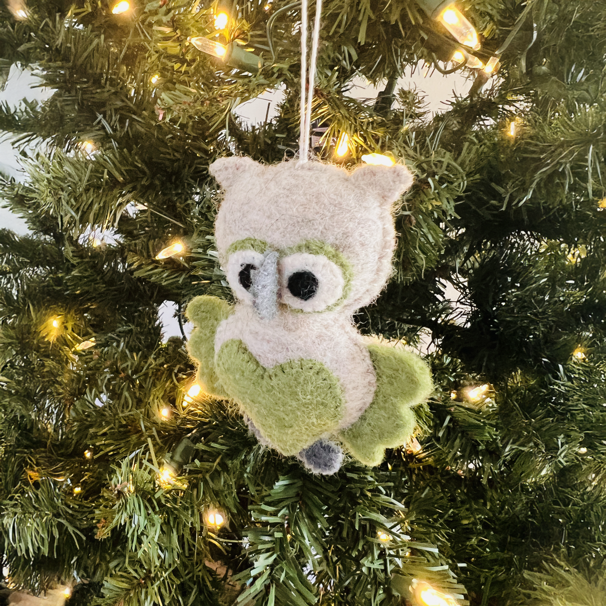 The Hand-Stitched Felted Owl Ornament featuring a green heart hangs on a lit Christmas tree, bringing charming detail to your artisan holiday ornament collection.