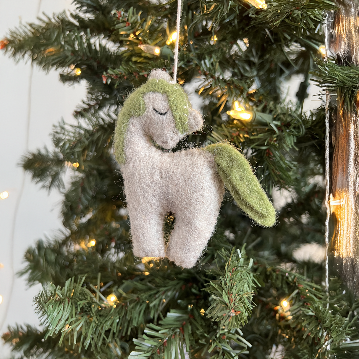 A Hand-Stitched Felted Small Unicorn Ornament with a green mane and tail hangs on a Christmas tree with lights, bringing unique charm to your artisan holiday ornament collection.