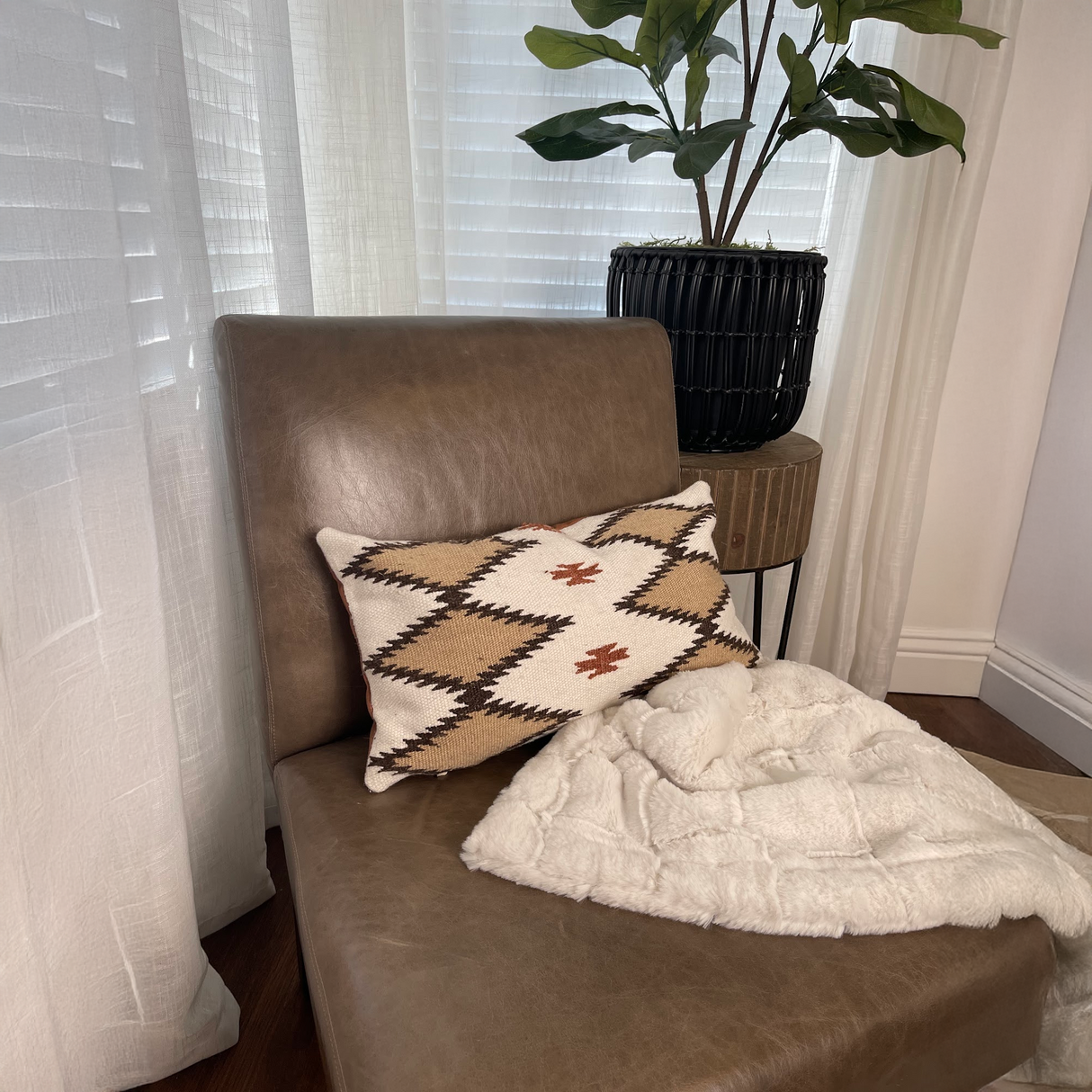 Brown leather chair with a Handwoven Blazheny (Blissful) Pillow, white throw, and a potted plant in a cozy corner by the window.