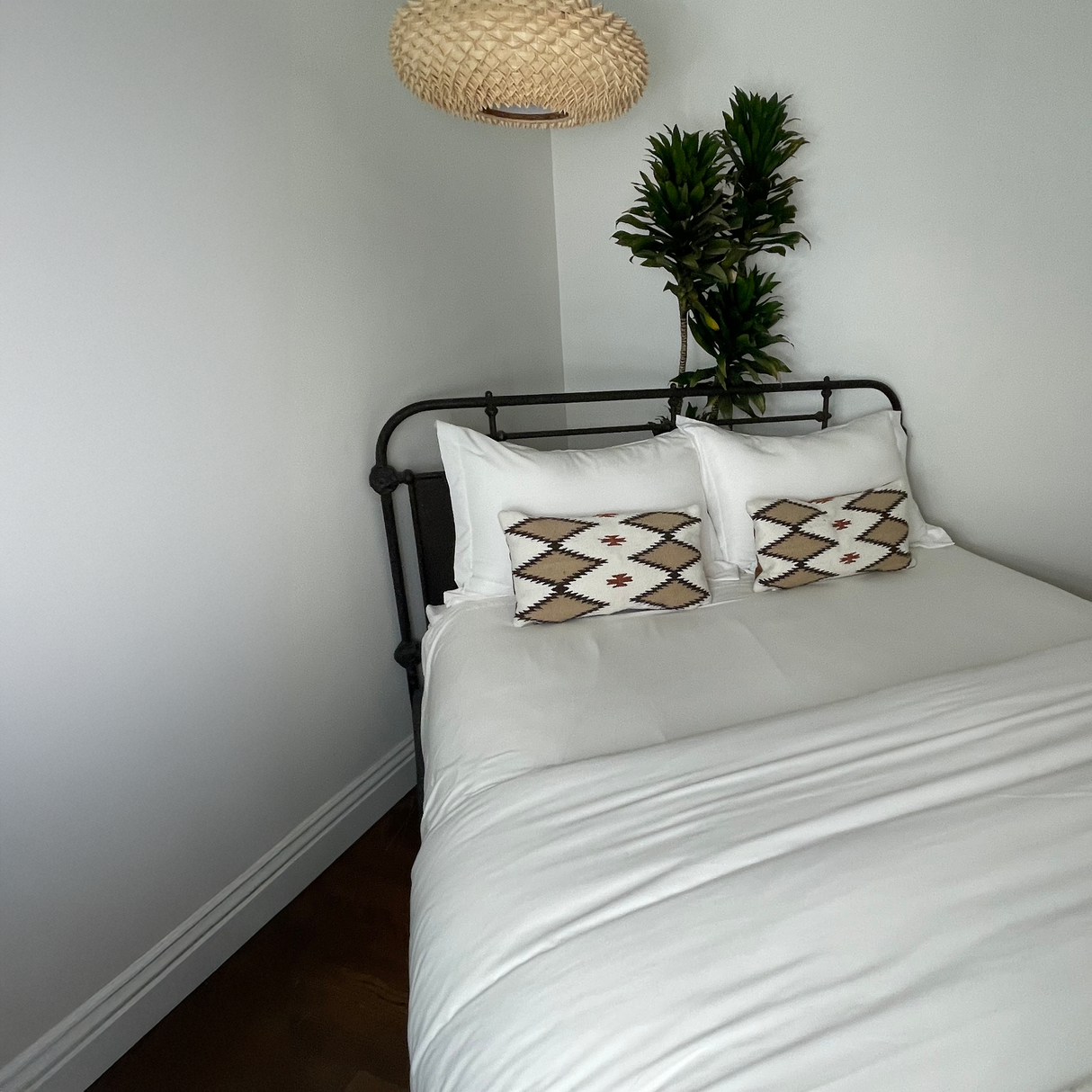 A neatly made bed with white linens, patterned pillows including the Handwoven Blazheny (Blissful) Pillow, a potted plant, and a woven pendant lamp above.