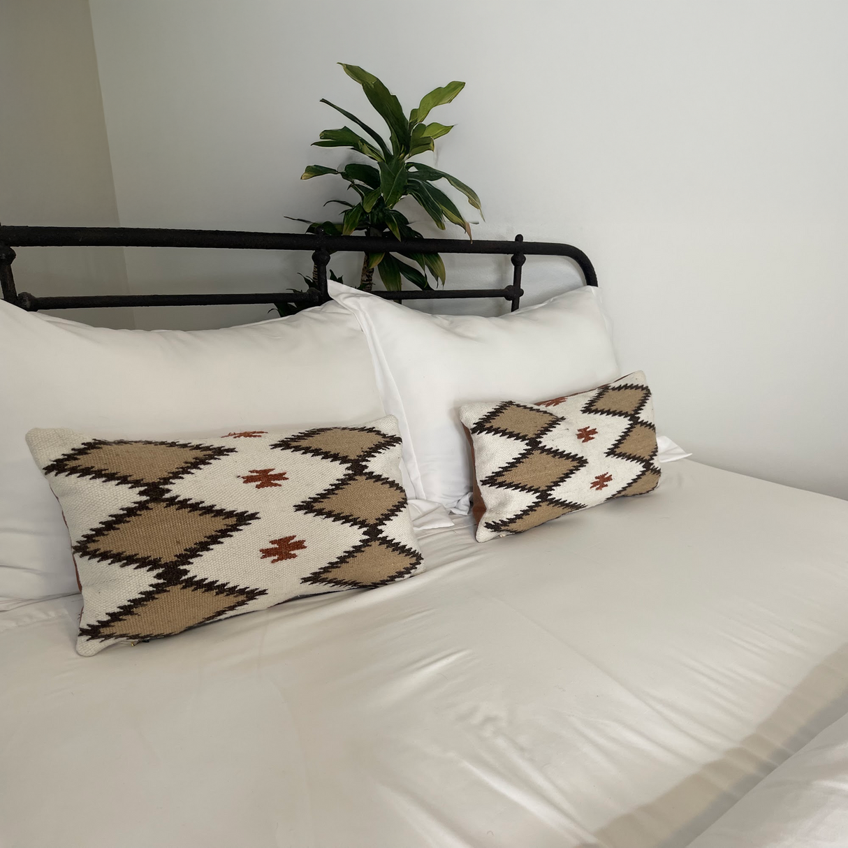 White bed with two patterned pillows, white bedding, and a green plant against a white wall. The Handwoven Blazheny (Blissful) Pillow adds artisanal charm to this serene setting.