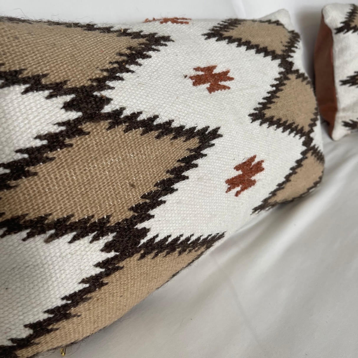 The Handwoven Blazheny (Blissful) Pillow, featuring a beige, white, and brown geometric design with small orange accents, is displayed on a white surface.