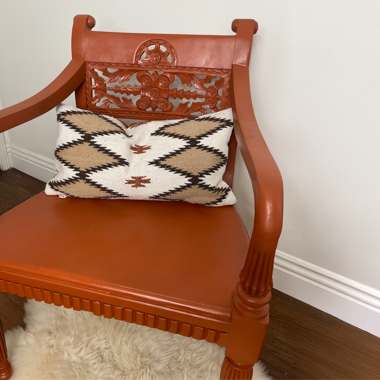 A carved wooden chair with an orange finish and patterned cushion is paired with the Handwoven Blazheny (Blissful) Pillow, set on a wood floor with a white rug.