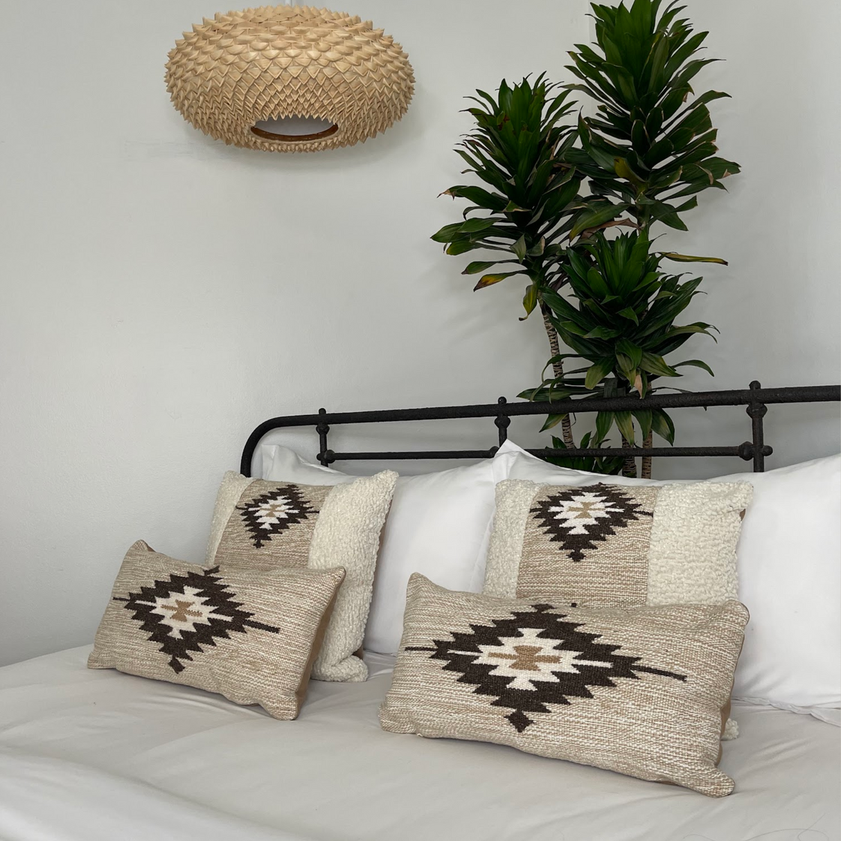 A neatly made bed with patterned pillows, the Handwoven Chitki (Crisp) Pillow, a tall plant, and a woven light fixture on the wall.