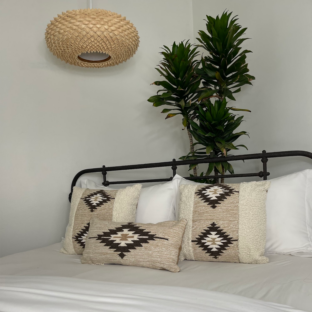 A minimalist room featuring a bed with the Handwoven Chitki (Crisp) Pillow, patterned cushions, a tall green plant, and a woven pendant light.