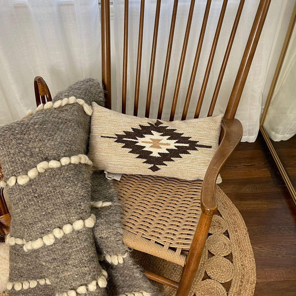 A wooden rocking chair with a Handwoven Chitki (Crisp) Pillow and textured blanket sits on a woven rug by a white curtain.
