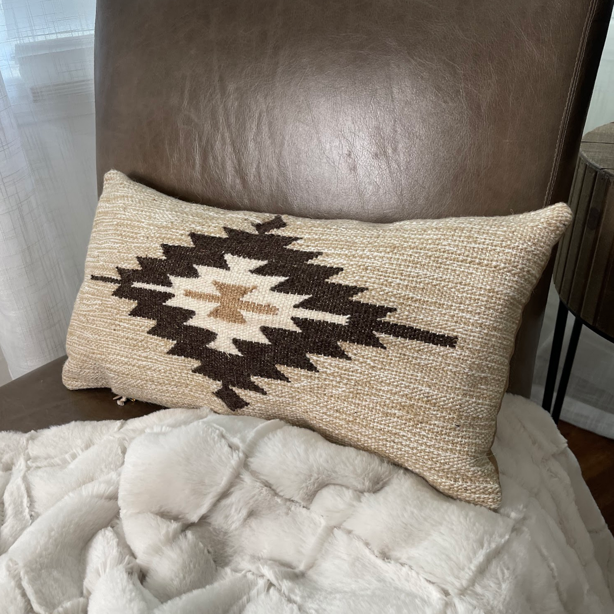 The Handwoven Chitki (Crisp) Pillow, featuring a brown geometric design, rests on a vegan leather chair draped with a white faux fur blanket.