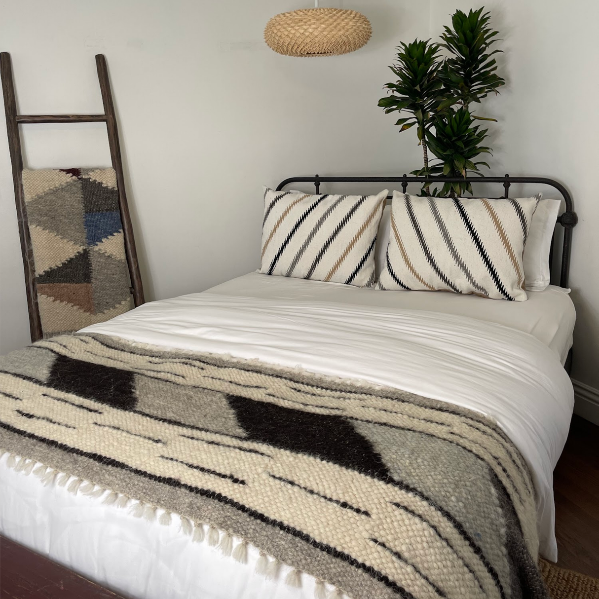 A neatly made bed features the Handwoven Promin (Beam) Wool Pillow, striped pillows, a patterned blanket, and plant and ladder decor beside it.