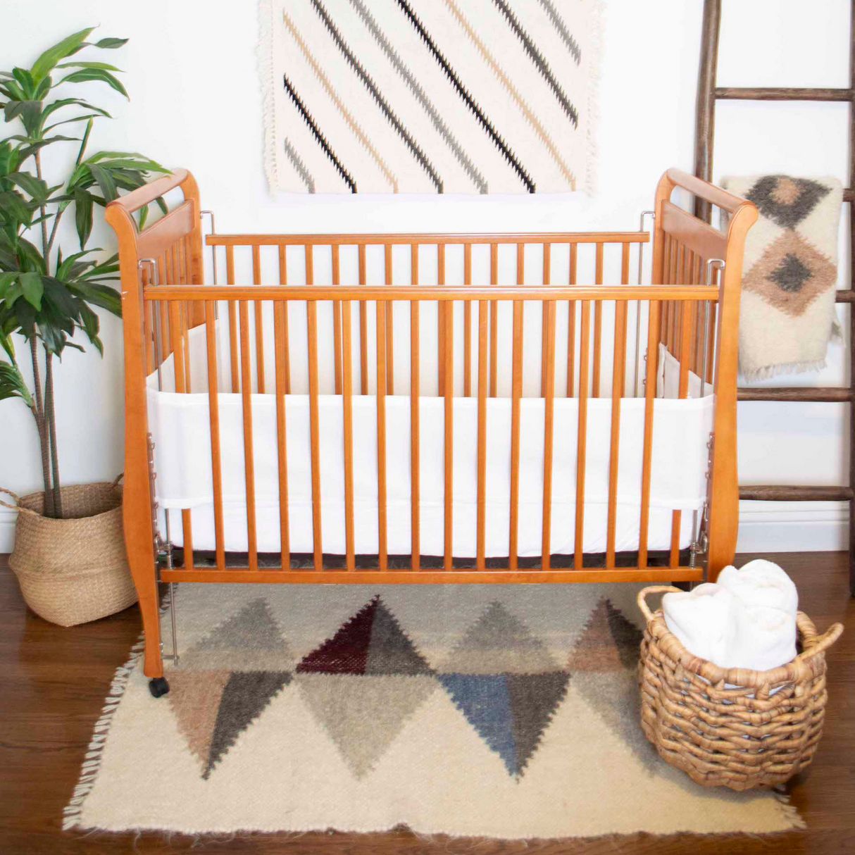 Wooden baby crib with a white bumper, Harmonia A (Harmony) Wool Statement Throw / Baby Blanket, geometric rug, plant, and basket of towels in a cozy nursery.