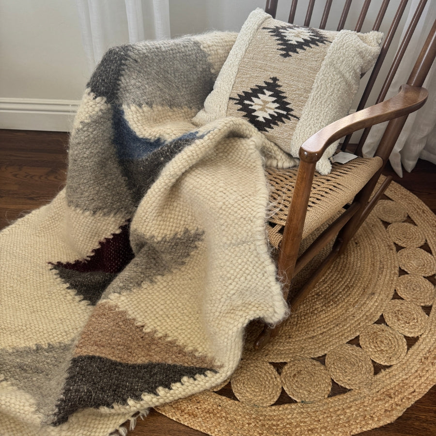 Wooden rocking chair with a woven seat, draped with the Harmonia A (Harmony) Wool Statement Throw / Baby Blanket and a patterned pillow, placed on a braided rug.