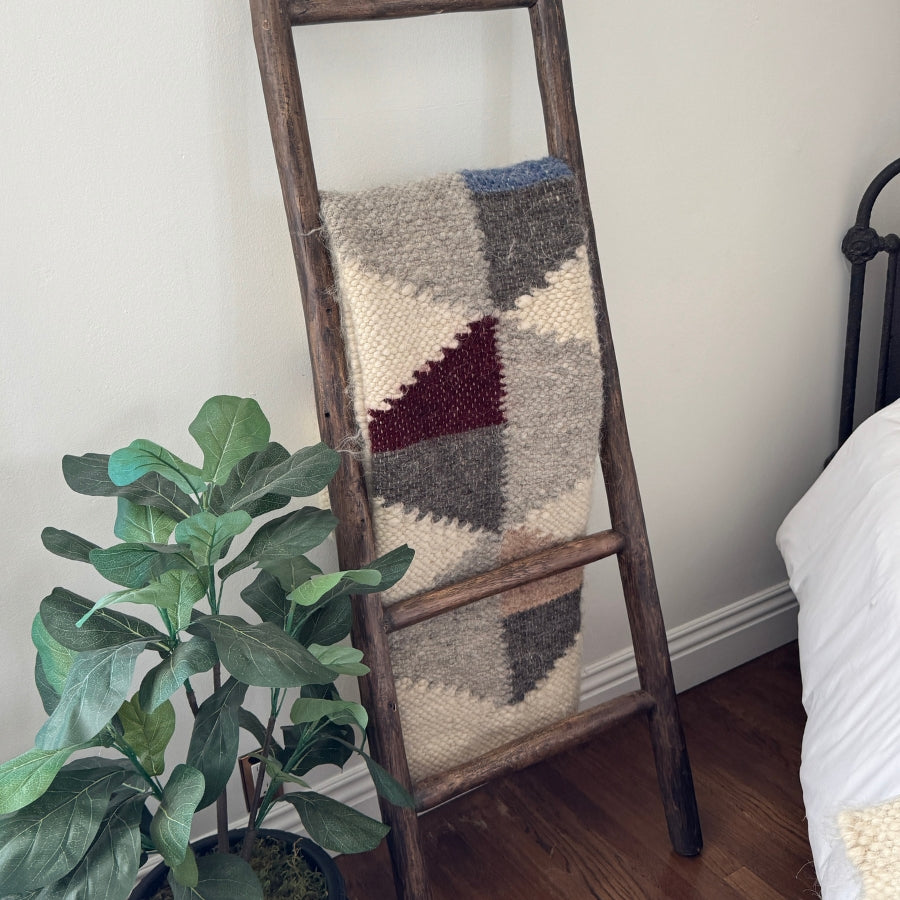 The Harmonia A (Harmony) Wool Statement Throw / Baby Blanket with a geometric-knit design is draped over a wooden ladder beside a green potted plant and a bed.