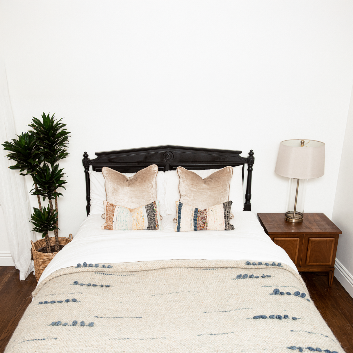 A neatly made bed with two pillows, the Horoshy (Kind) Wool Statement Bed Blanket crafted by Ukrainian weavers, a potted plant, and a lamp on a wooden nightstand.