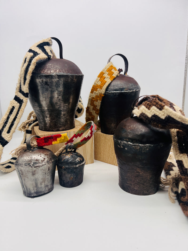 Five rustic metal cowbells with colorful woven straps displayed on light wooden blocks against a white background.
