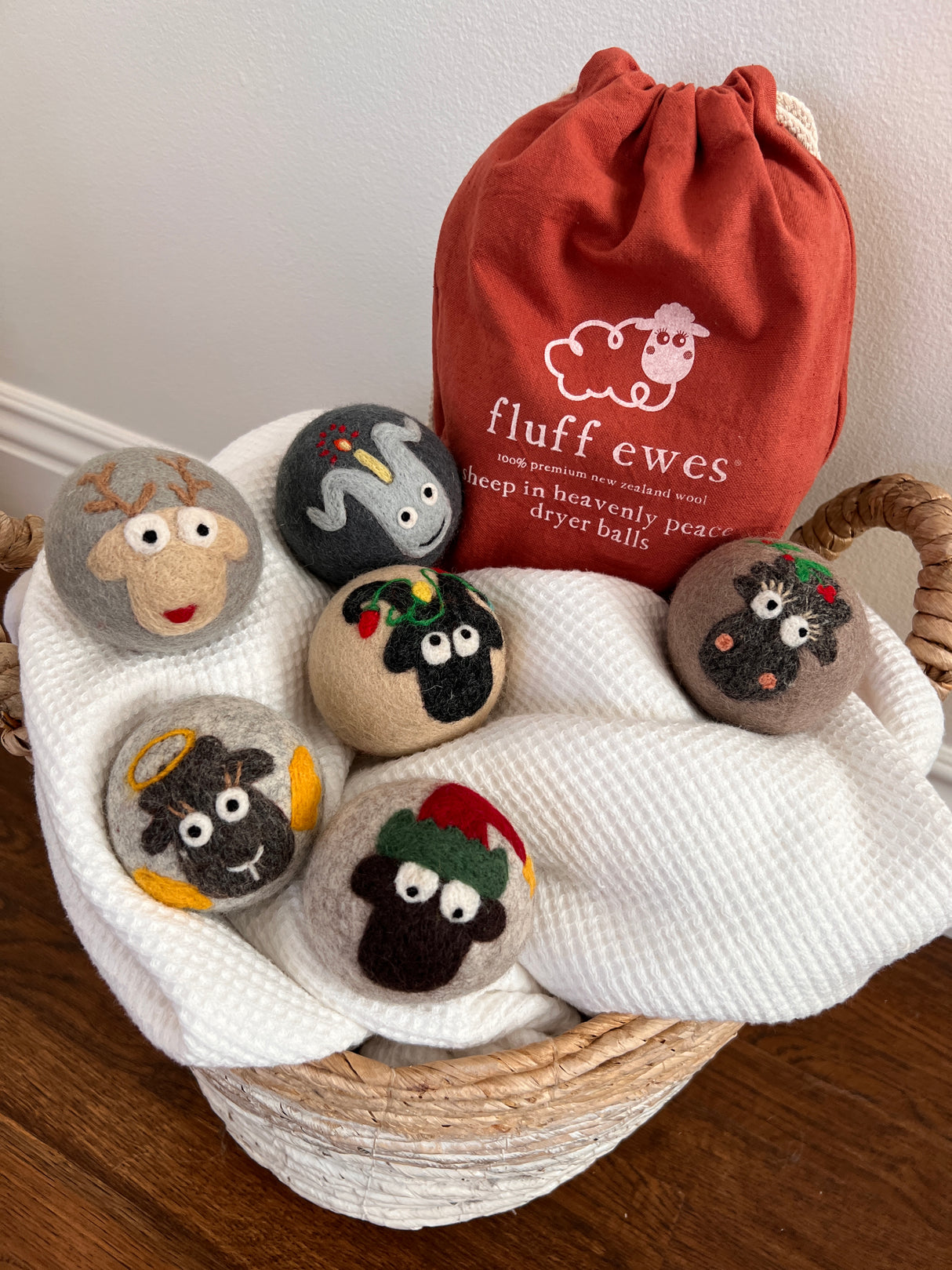 The Fluff Ewes Premium XL Dryer Balls "Sheep in Heavenly Peace" Holiday Limited Edition 6-pack features eco-friendly, animal-themed wool dryer balls in a red "fluff ewes" bag, all presented in a basket lined with white cloth.