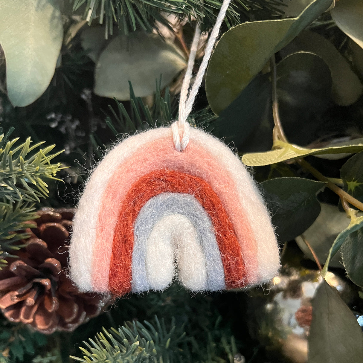 Rainbow is a hand-felted ornament crafted from New Zealand wool, featuring pink, red, and gray arches. Hang this artisan Rainbow on your Christmas tree for festive décor with a touch of greenery and pinecones.