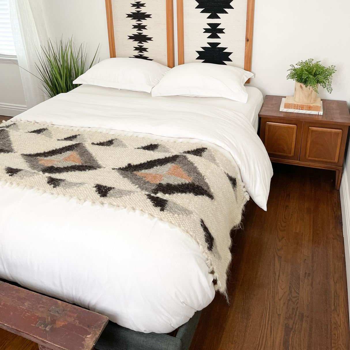 Minimalist bedroom with a white bed, geometric blanket, wooden nightstand, plants, and a hardwood floor, featuring the Iskara (Spark) Wool Bed / Couch Runner for extra warmth and texture.
