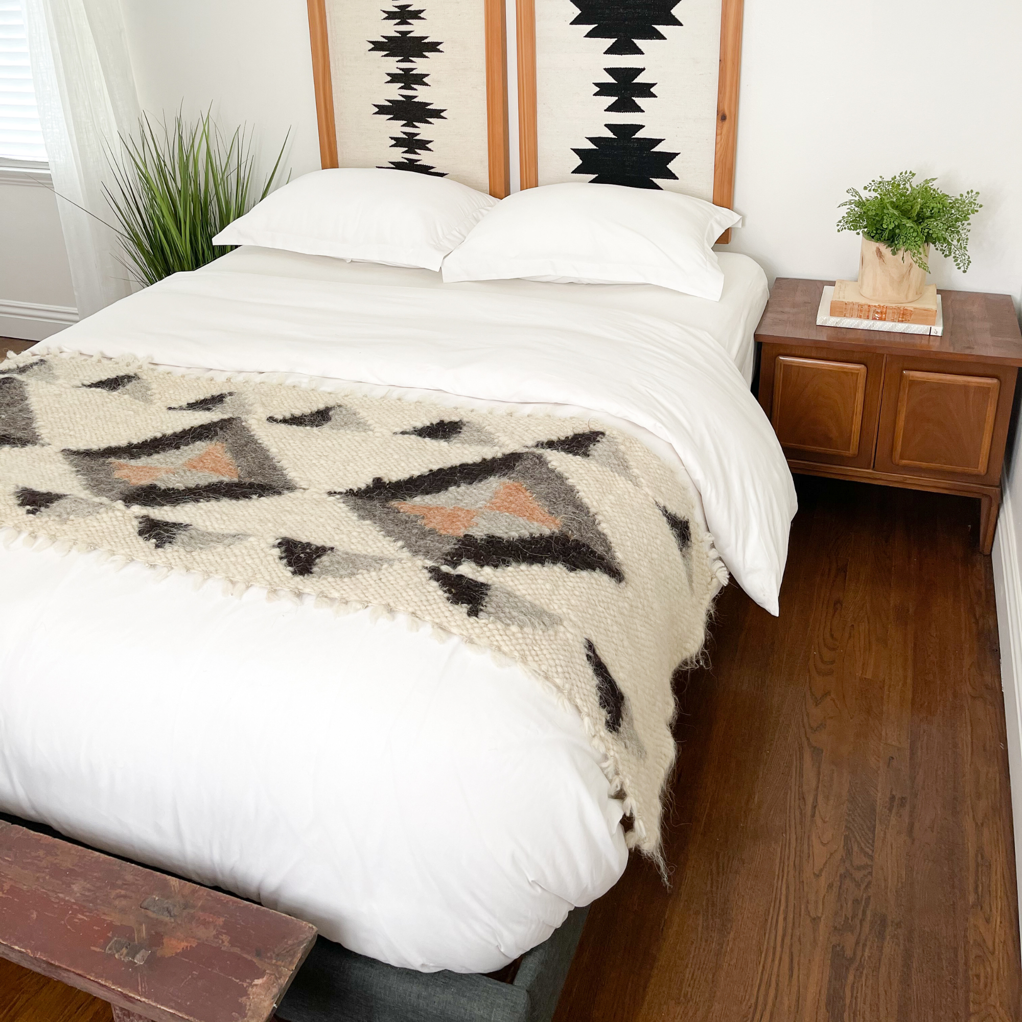Minimalist bedroom with a white bed, geometric blanket, wooden nightstand, plants, and a hardwood floor, featuring the Iskara (Spark) Wool Bed / Couch Runner for extra warmth and texture.