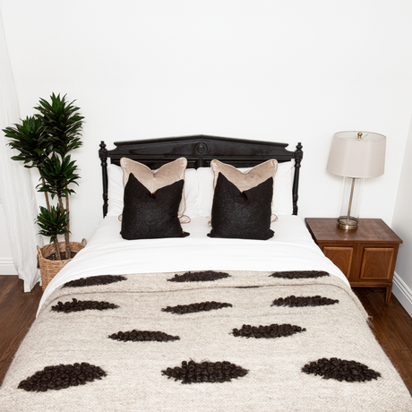 A neatly made bed with two black pillows, the Khmara (Cloud) Wool Statement Bed Blanket, a plant, and a lamp on a nightstand.