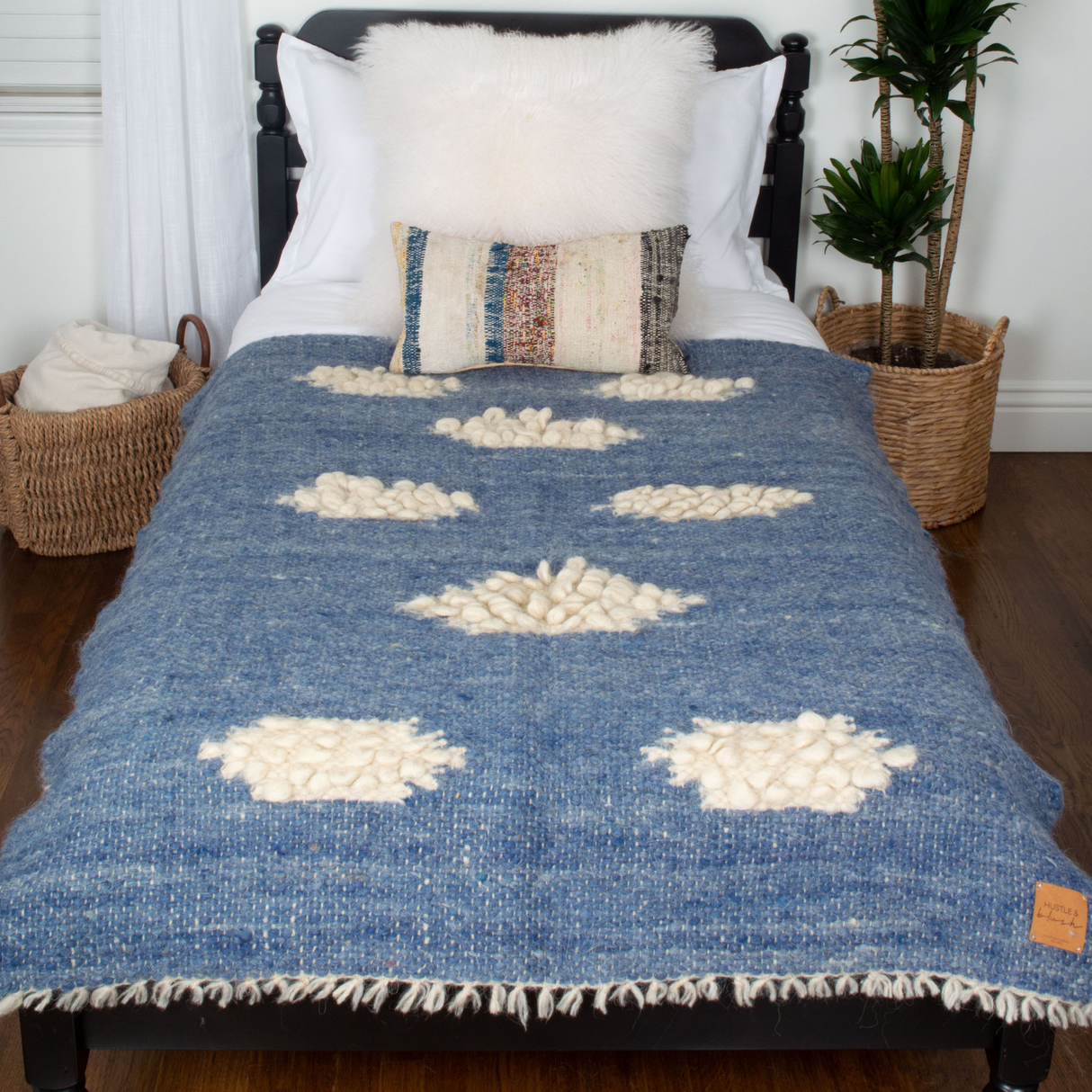 A bed with the Khmara (Cloud) Wool Statement Throw featuring white cloud patterns, a fluffy pillow, and baskets beside it on a wooden floor.