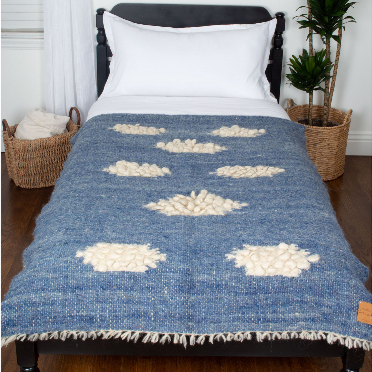 The Khmara (Cloud) Wool Statement Throw / Baby Blanket, in blue and white tufted wool, adds texture to a twin bed, complemented by nearby woven baskets and plants.