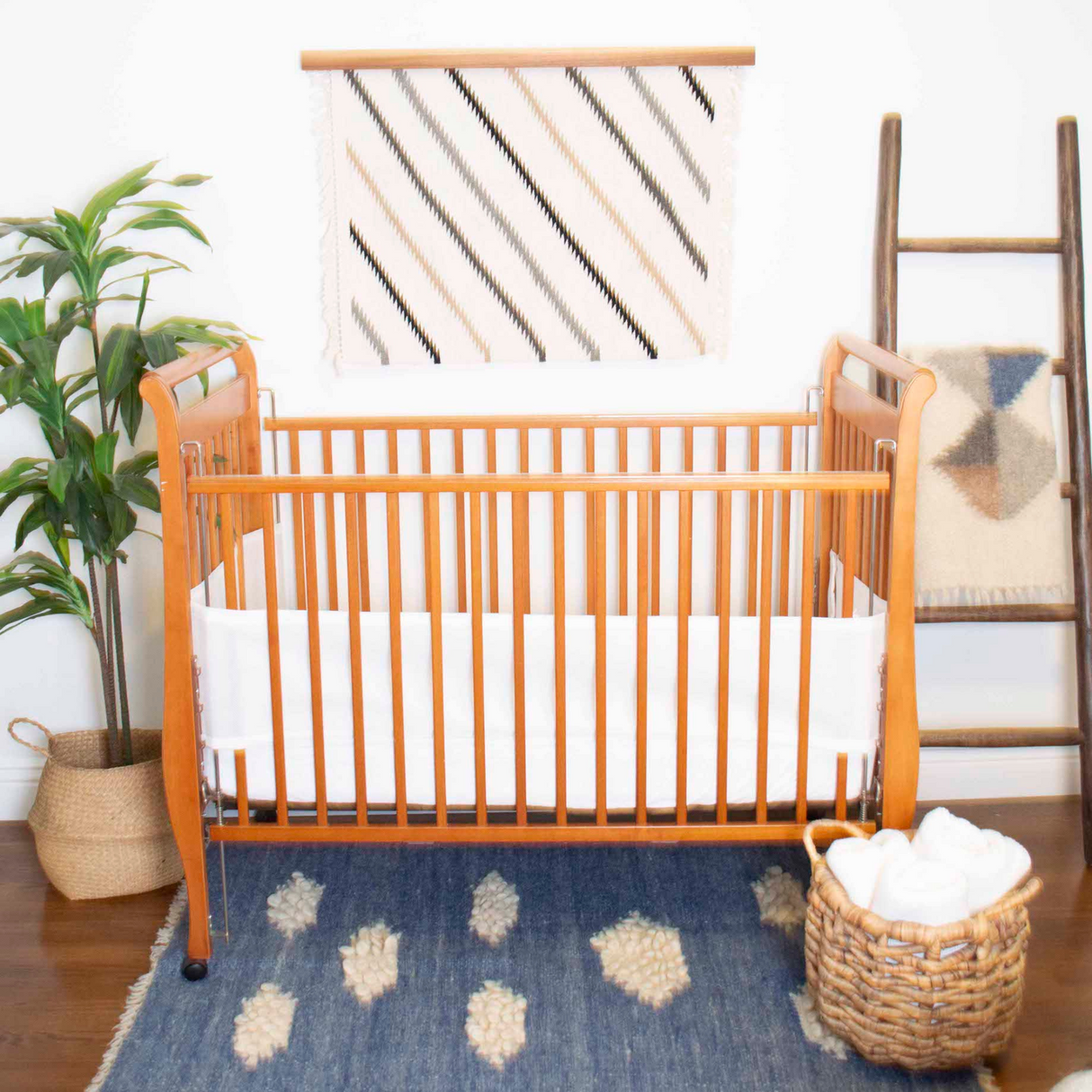 A natural wood crib with white bedding and the Khmara (Cloud) Wool Statement Throw adds warmth, while a plant, woven basket, ladder, and blue rug complete this cozy, inviting nursery space.