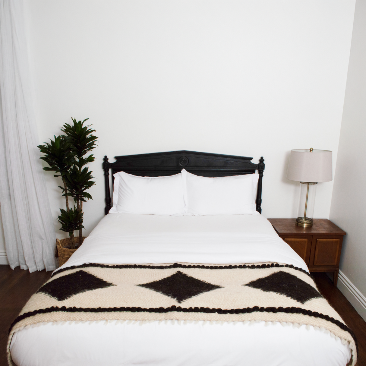 A neatly made bed with white linens, a Korysno Wool Bed/Couch Runner of 100% virgin wool, a lamp, and a potted plant beside it.