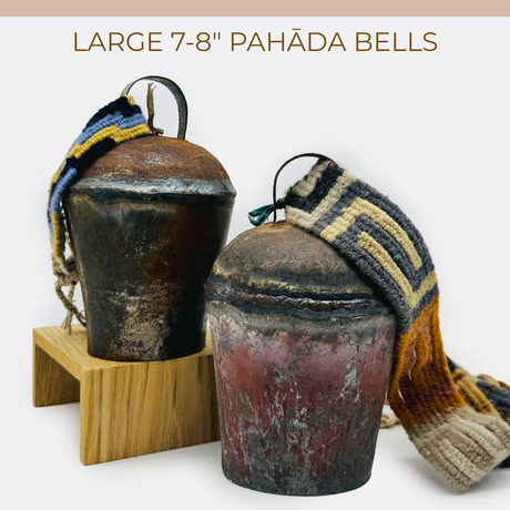 Two vintage handmade Pahāḍa yak, goat & sheep’s bells (7-8") from the Himalayan mountains of Nepal and Tibet, with colorful woven straps, rest on a wooden stand—perfect as Christmas ornaments, wreath décor, or for sound baths.