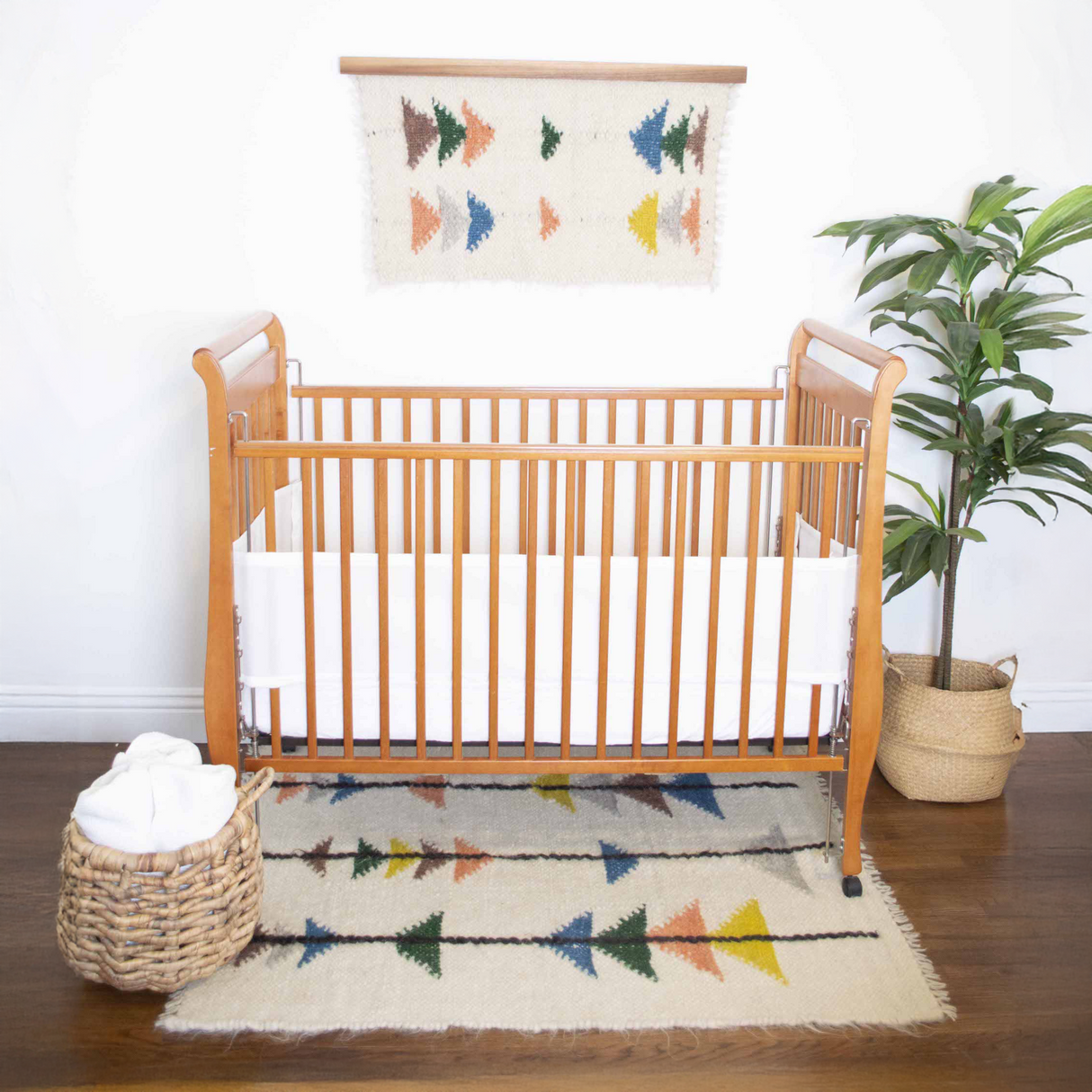 A wooden crib with a white sheet, the Lis (Forest) Wool Statement Throw / Baby Blanket, a plant in a basket, and a colorful geometric rug create a welcoming nursery.