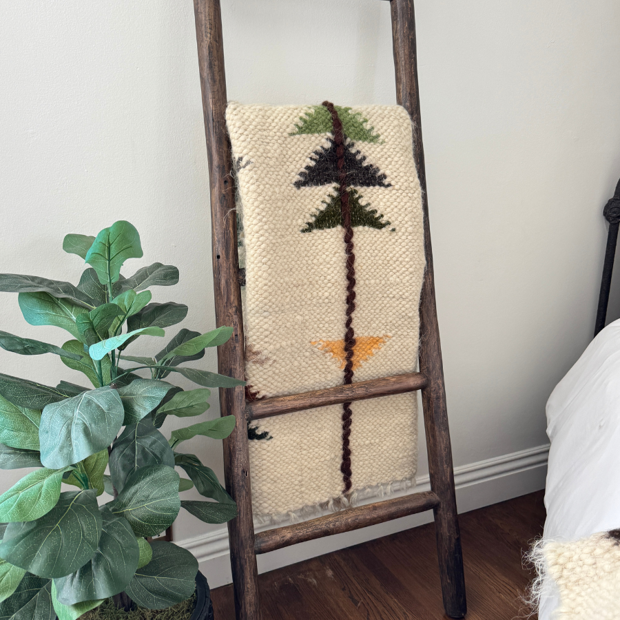 The Lis (Forest) Wool Statement Throw / Baby Blanket with geometric patterns hangs on a wooden ladder beside a potted plant in a cozy room.