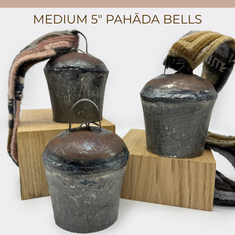 Three vintage handmade 5" Pahāḍa yak, goat, and sheep’s bells from the Himalayas of Nepal and Tibet, each with fabric straps, are displayed on wooden blocks. Ethically sourced fair trade metal bells perfect for decor or sound baths.