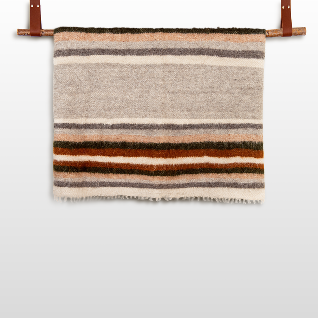 The Mudry (Wise) Wool Statement Baby Blanket, featuring brown, gray, orange, and cream stripes, hangs on a rod with leather straps.