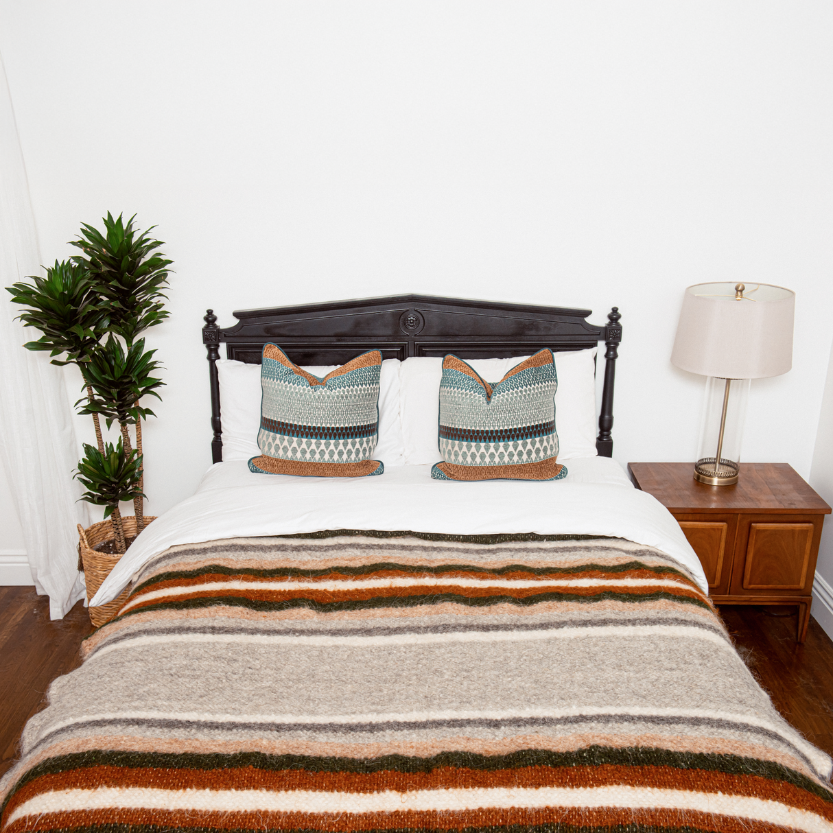 A neatly made bed with patterned pillows, the Mudry (Wise) Wool Statement Bed Blanket, a plant, and a lamp on a wooden nightstand.