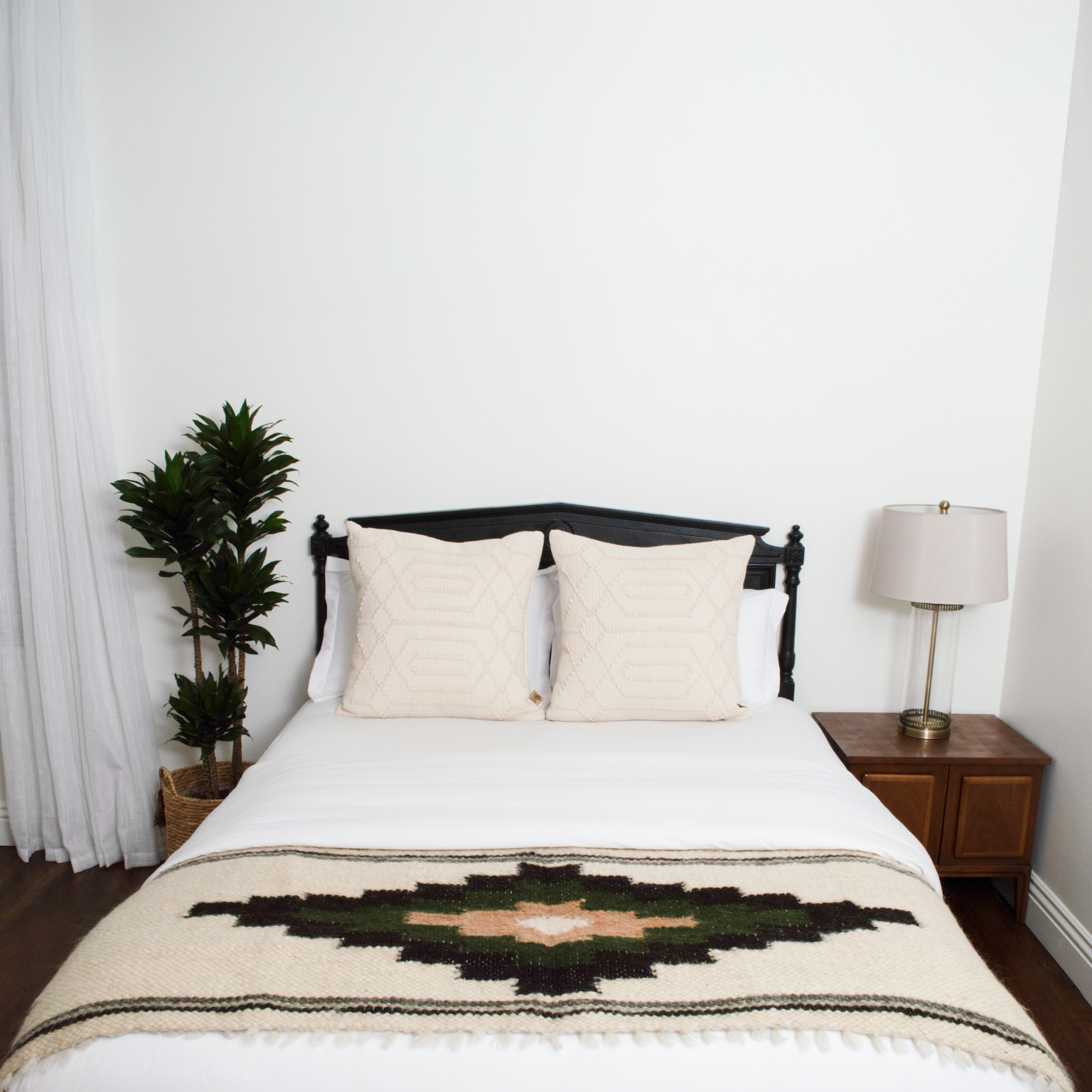 A minimal bedroom featuring a neatly made bed with the Myr (Peace) Wool Bed / Couch Runner, two pillows, a lamp, and a potted plant.