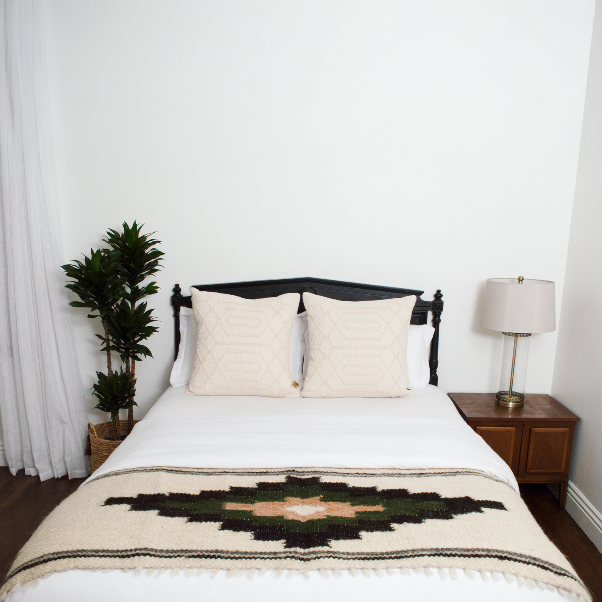 A minimal bedroom featuring a neatly made bed with the Myr (Peace) Wool Bed / Couch Runner, two pillows, a lamp, and a potted plant.