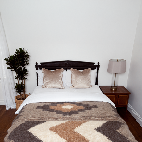 A neatly made bed with beige pillows, the Myr (Peace) Wool Statement Bed Blanket, a bedside table, lamp, and a potted plant.