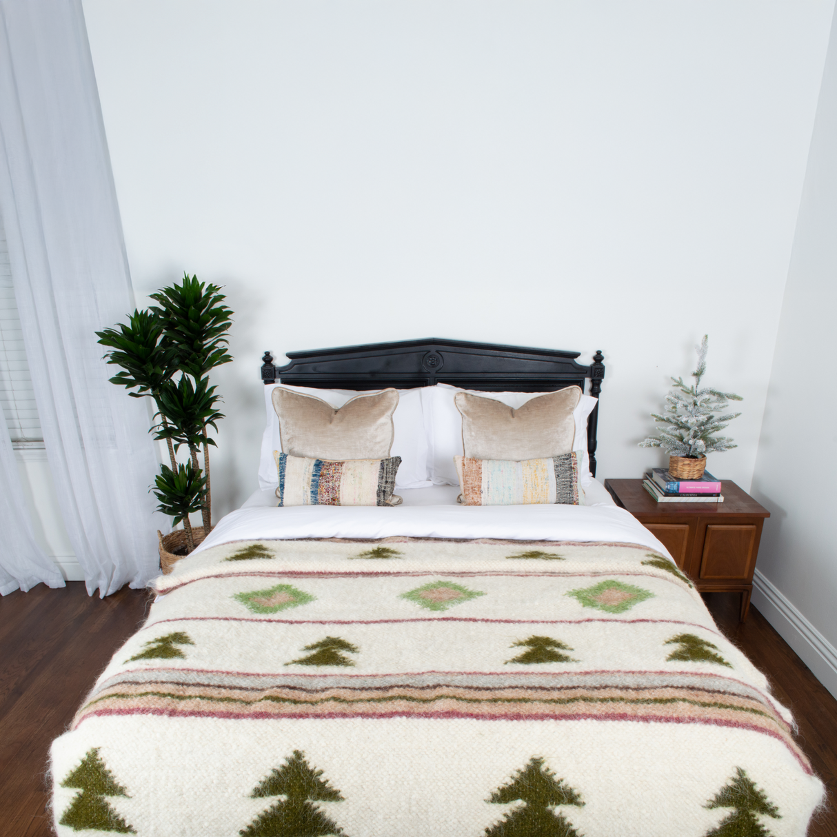 Bed with a Sosna (Pine) Wool Statement Bed Blanket, two pillows, plants, and a small tree on the bedside table, set against a white wall.