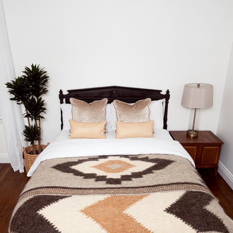 A neatly made bed with beige pillows, the Myr (Peace) Wool Statement Bed Blanket, a potted plant, and a lamp on a nightstand.
