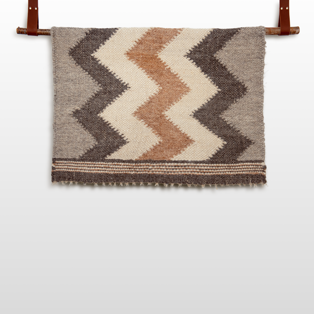 The Myr (Peace) Wool Statement Throw / Baby Blanket, made from all-natural undyed yarn, displays brown, tan, and cream chevron patterns and elegantly hangs on a wooden rod with leather straps.