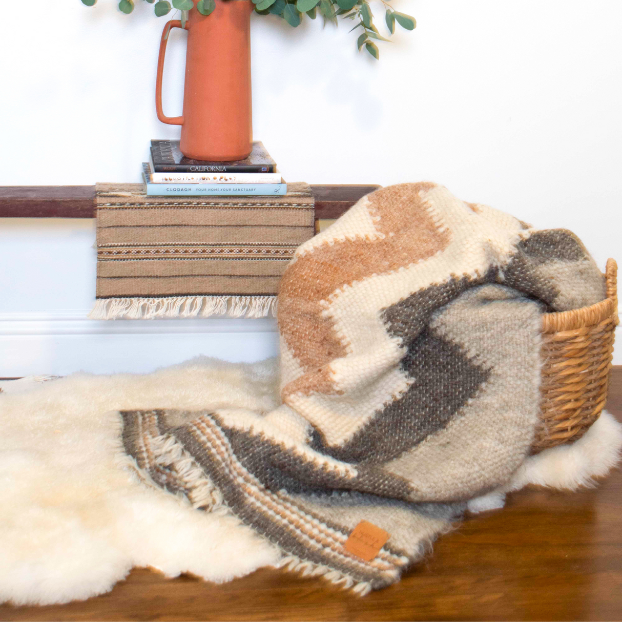 The Myr (Peace) Wool Statement Throw / Baby Blanket rests in a basket on a fur rug, with a rust-colored pitcher and a woven mat adding warmth and texture to the background.