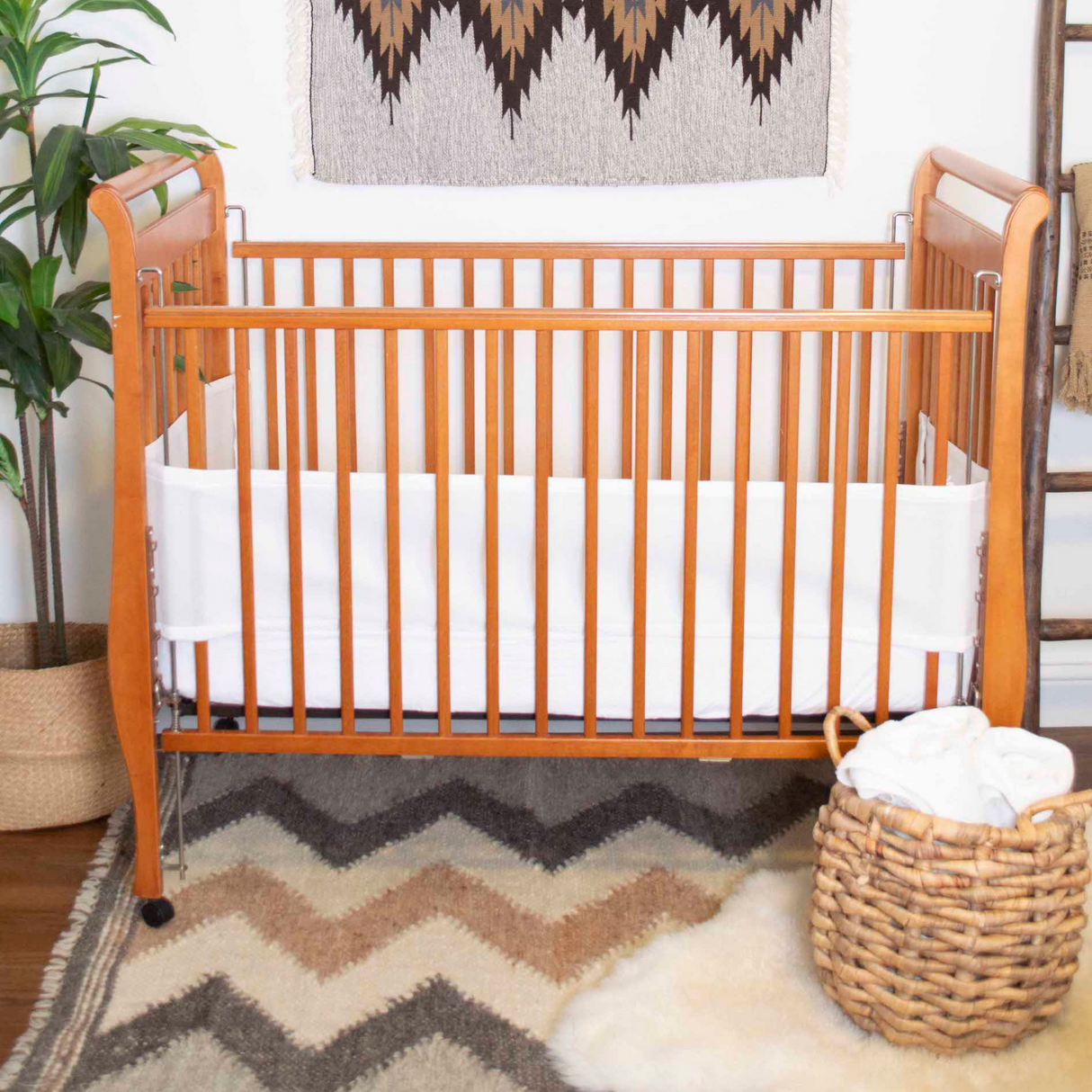 Wooden baby crib with white lining sits on a chevron rug beside a basket, green plant, and the Myr (Peace) Wool Statement Throw / Baby Blanket for extra comfort.