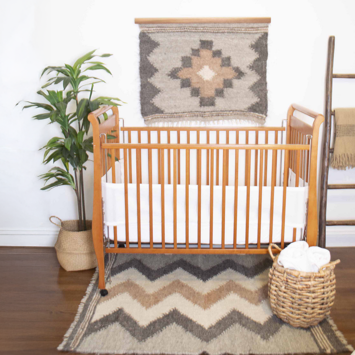 A wooden crib on a patterned rug, with a plant, basket, and the Myr (Peace) Wool Wall Hanging made from all-natural undyed yarn, adds warmth and traditional Ukrainian design to this cozy nursery.