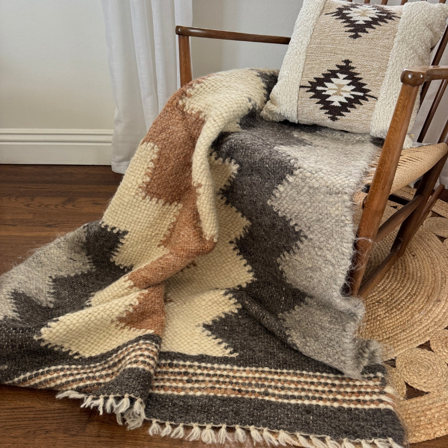 A wooden chair with a woven pillow and the Myr (Peace) Wool Statement Throw—crafted from all-natural undyed yarn—draped over the arm, adds cozy charm.