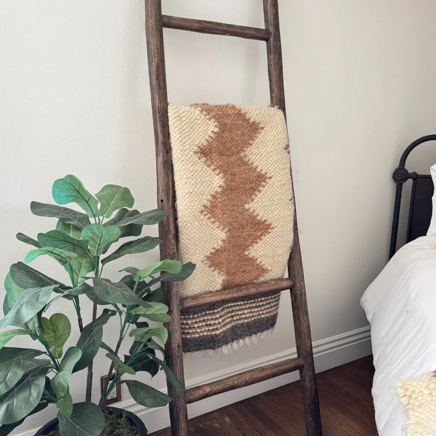The Myr (Peace) Wool Statement Throw / Baby Blanket is draped over a wooden ladder beside a plant and a white bed in a cozy room.