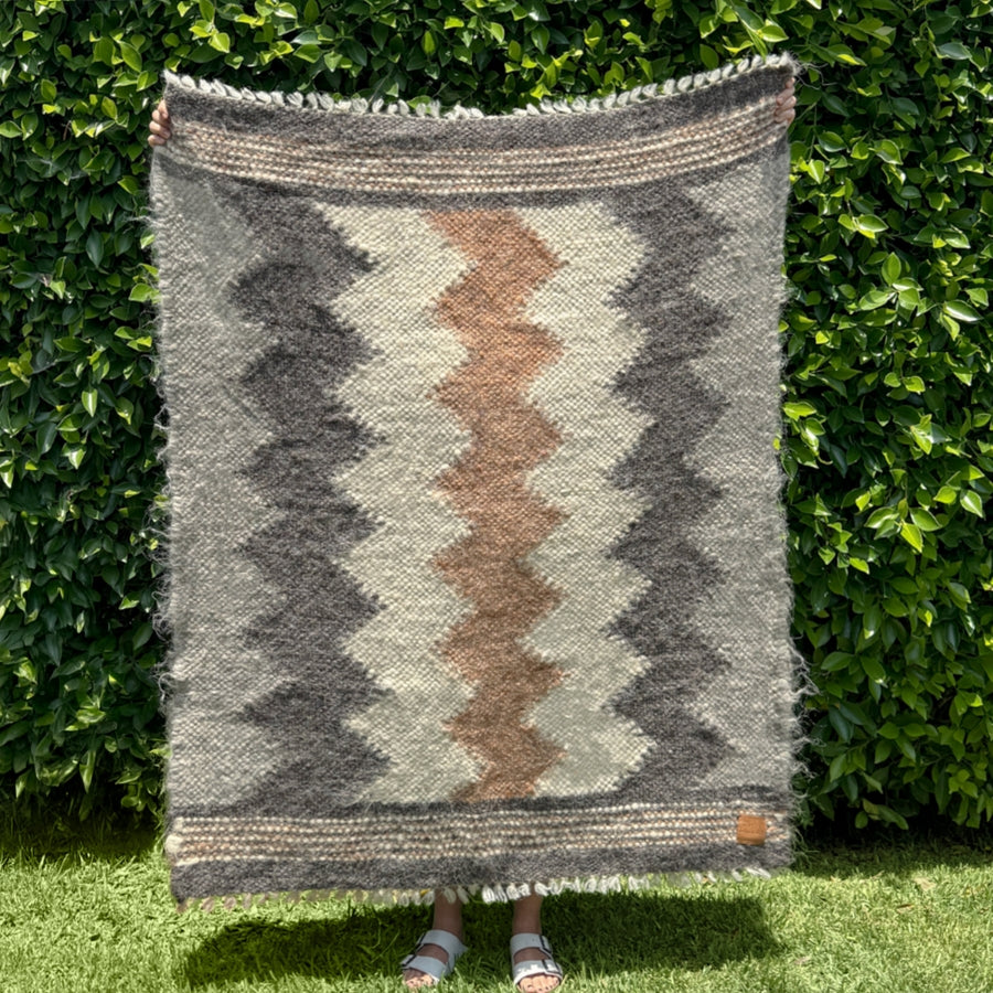 Person holding the Myr (Peace) Wool Statement Throw / Baby Blanket, featuring brown, gray, and cream chevron zigzags in all-natural, undyed yarn, standing before green hedges.